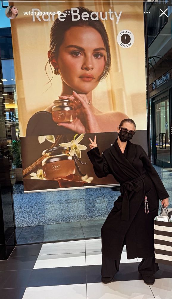 Selena Gomez wearing a black mask and sunglasses in front of a photo advertisement featuring herself.