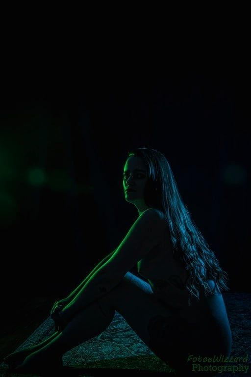 Young woman sitting nude in a dark room on a rug, rim lit with green and blue light. 