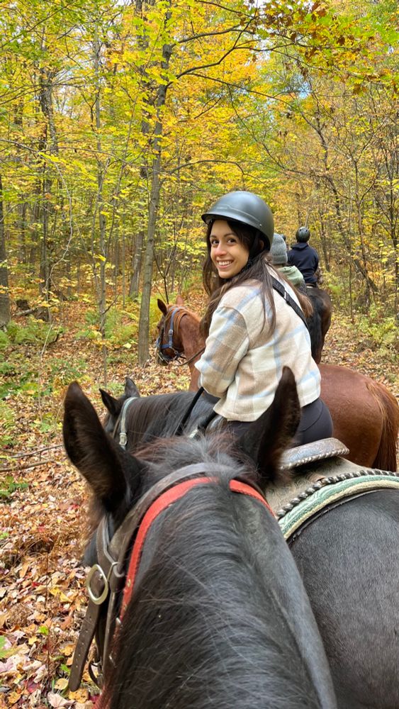 me (caucasian brunette) horseback riding with fall foliage all around me. I’m wearing a beige and blue plaid shacket, black helmet, smiling back at the camera. I’m on a black horse and another horse is also in the foreground. 