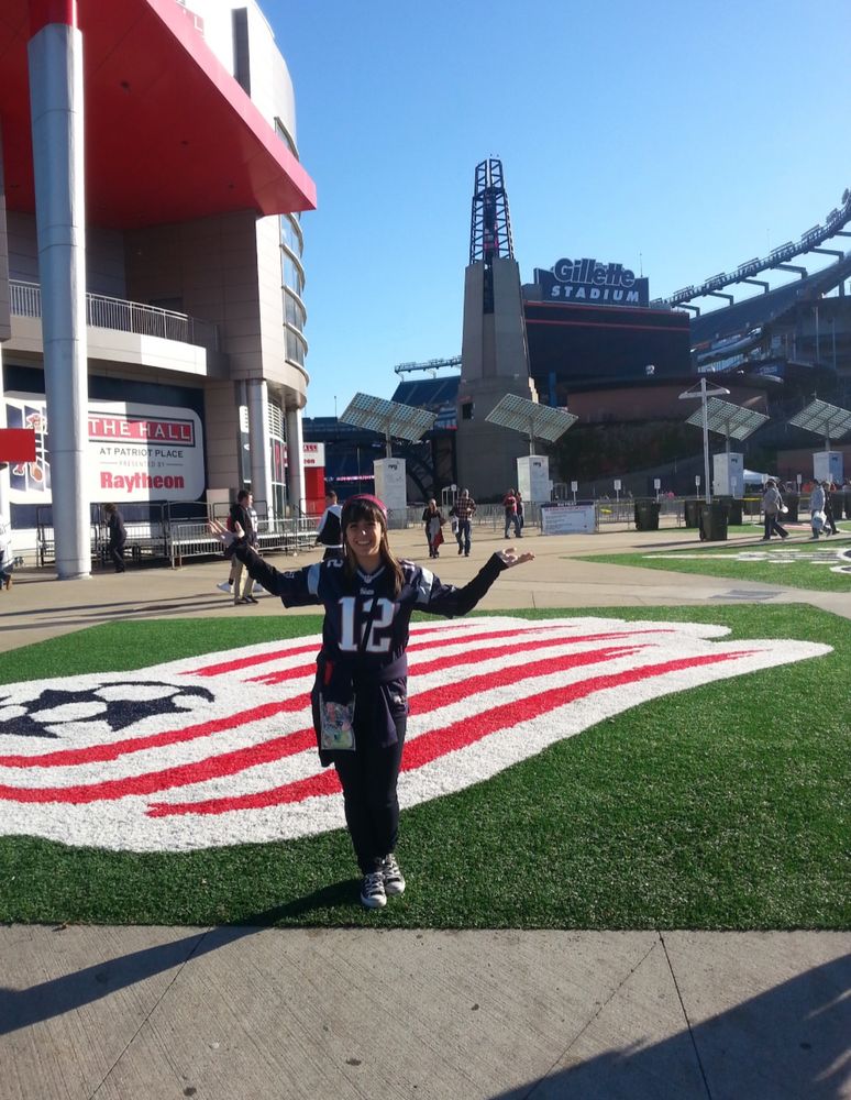 younger me with brown hair and 2 blonde streaks on each side with bags standing at Gillette stadium (green turf with NE soccer flag) wearing Patriots gear (Jersey number 12, headband, and purse) with my arms raised.