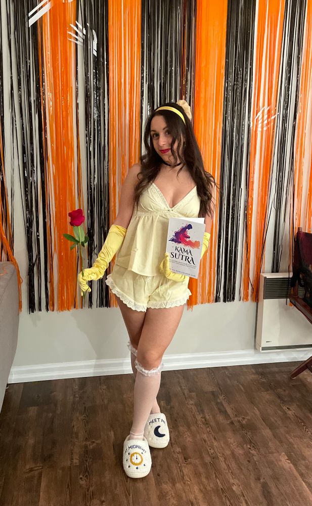 brunette woman (me) dressed in yellow lingerie with my hair like Belle’s, yellow gloves, white knee high socks, holding a rose and kama sutra book. I am wearing “meet me at midnight” slippers and posing in front of orange and black decor.
