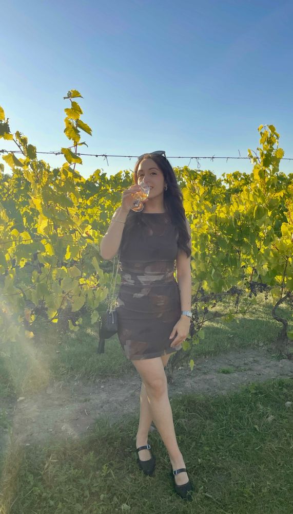 caucasian woman standing in a vineyard (lush greenery and vines behind her) holding a glass of white wine to her lips. she has long brown hair, wearing a brown floral patterned tight dress and black kitten heels and silver jewelry 