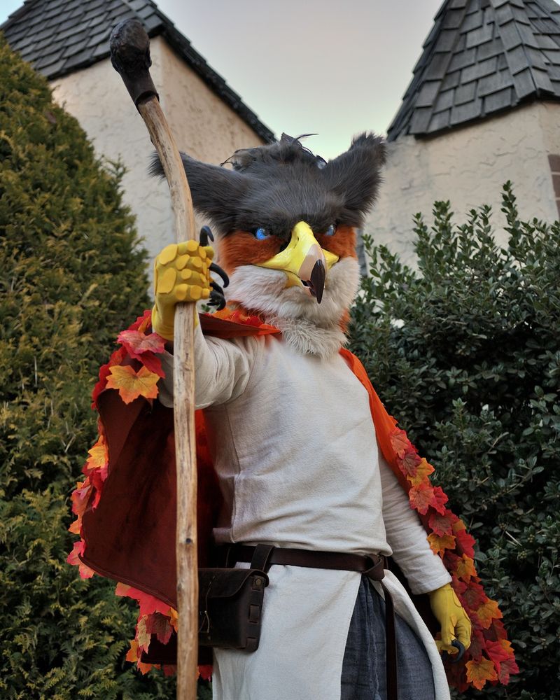 A gryphon partial fursuit with a head and gloves. He's wearing a cape made of leaves and is carrying a wooden staff. 