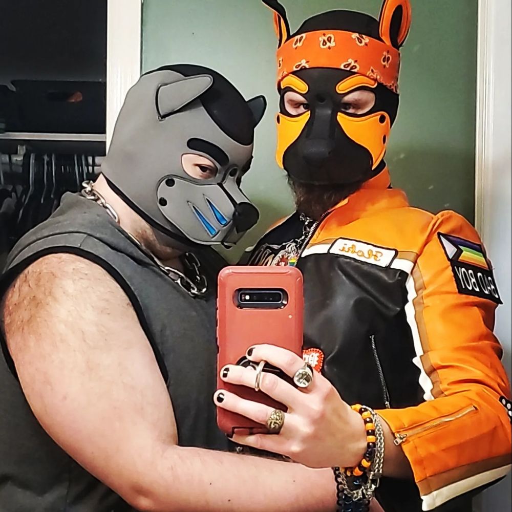 Artto and Floki, dressed in pup hoods and gear, stand side-by-side for a mirror selfie before going out for a pup night event.