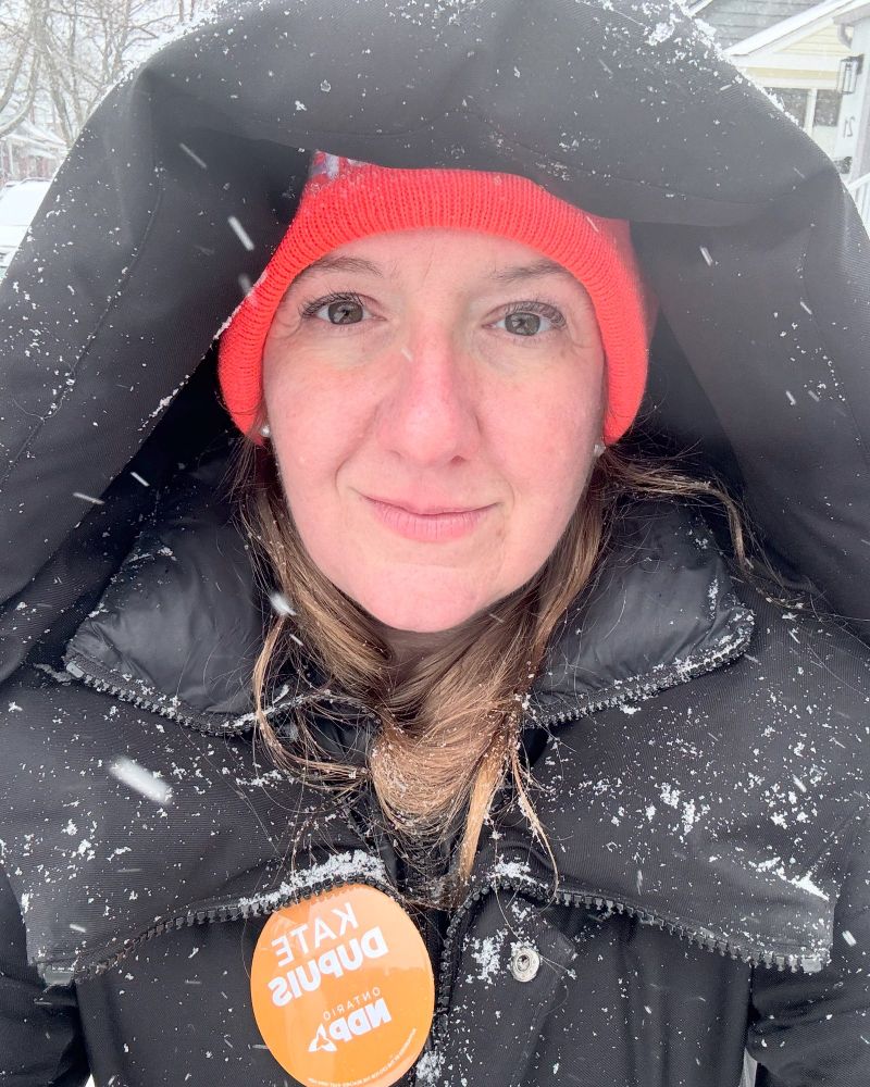 A woman stands in a black coat and orange toque. There is snow all over her coat and her hood is up over her head. 