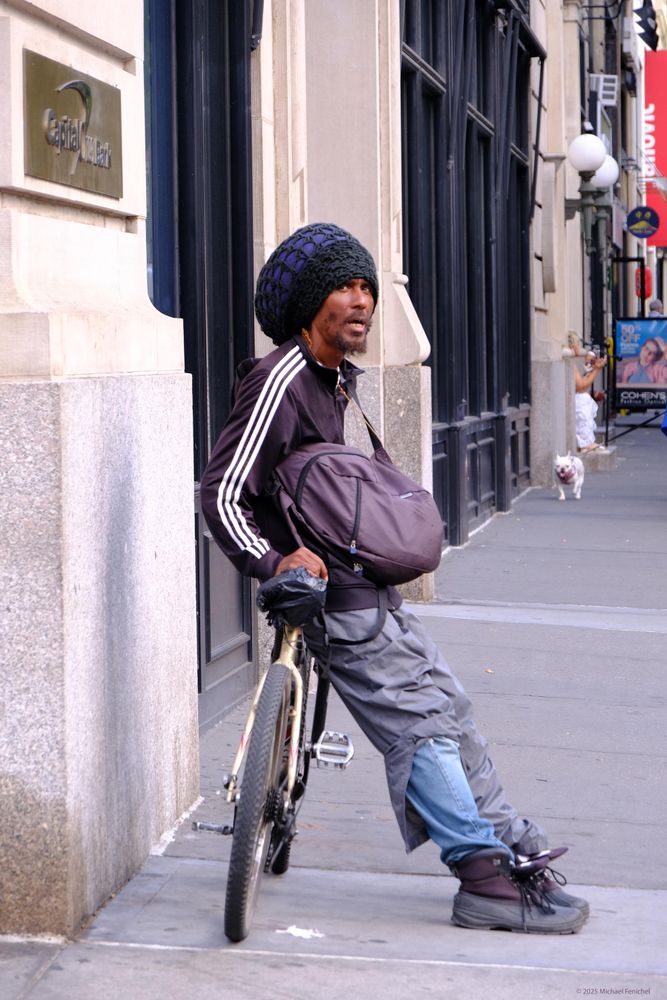 Photograph by Fenichel

A man is leaning back seated on a bicycle seat, but perpendicular to the bike, both feet planted on the ground, revealing some fancy footwear and (untied, high-top shoes of some sort) over jeans sheathed in another pair of cut-off pale purple pants. Looking upward he has slung over his shoulder, a purple bag with strap, a "Courier" style bag, and he wears a similarly colored striped arm jacket, and a wool hat covering perhaps a mountain of hair.

As the man is just relaxing, behind him a happy looking white dog is prancing past a doorway, in the direction of the bicyclist.