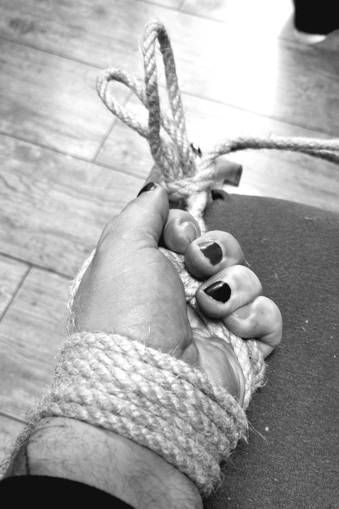 Schwarz-weiß Foto: Eine Männerhand ist mit geflochtenen Bondage Seilen an einem Bett festgemacht. Die Handfläche ist oben.