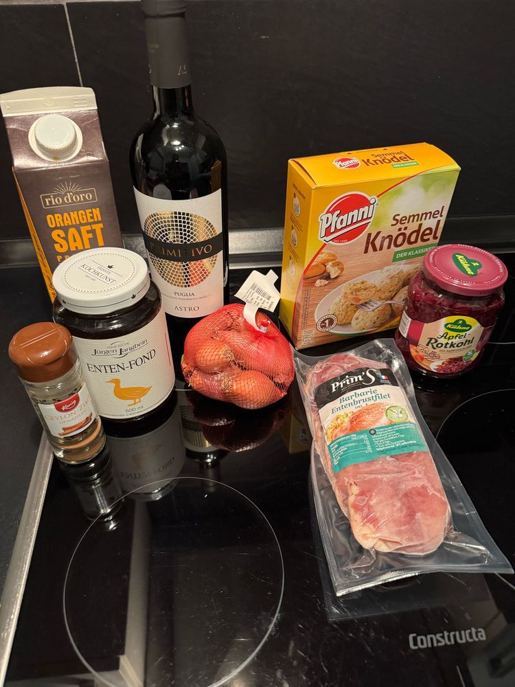 Das Foto zeigt eine noch verpackte rohe Barbarie-Entenbrust, noch verpackte Semmelknödel von Pfanni, ein Glas Apfelrotkohl von Kühne, ein Netz Schalotten, ein Glas Enten-Fond, eine Flasche Rotwein Primitivo), ein Packet Orangendirektsaft sowie einen Zimtstreuer - alles drapiert auf einem Ceranfeld. 