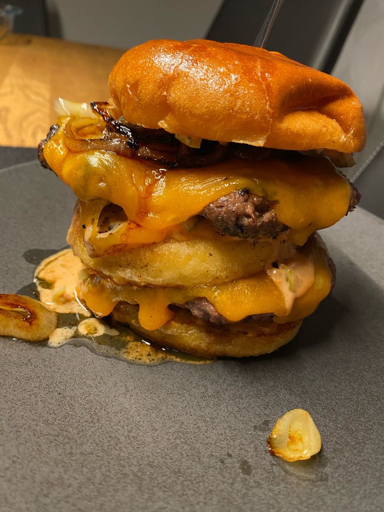 Ein vor Butter und Fett triefender Burger: 1. Bun, Soße, karamellisierte Zwiebeln, Patty mit Cheese, Gurkenscheiben, 2. Bun mit Soße, karamellisierte Zwiebeln, Patty mit Cheese, Gurkenscheiben, 3. Bun mit Soße.