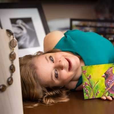 Image of Katherine Klimitas laying down on her side smiling in a green sleeveless dress while holding a painting of a cardinal that she painted.