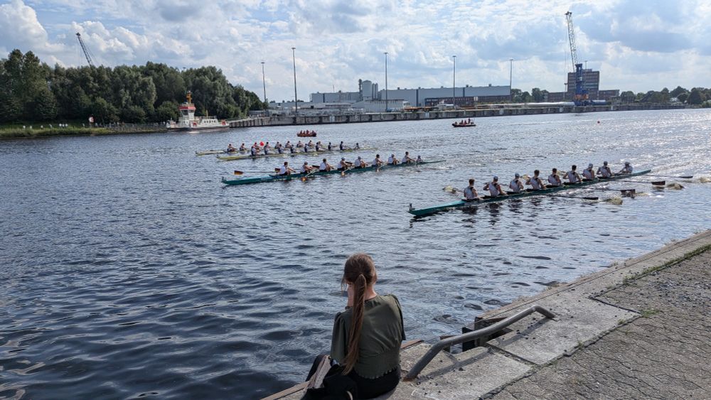 4 Achter auf dem Kanal, im Vordergrund eine Person auf der Kaimauer, im Hintergrund Begleitboote, eine Fähre und das andere Ufer mit Hafenanlagen und Bäumen