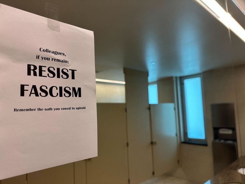 The image shows a printed sign taped to a mirror in what appears to be a public or office restroom. The sign is made from a standard sheet of white paper with black printed text. It is affixed at the top with a single piece of transparent tape.

The text on the sign reads:

⸻

Colleagues,
if you remain:
RESIST
FASCISM
Remember the oath you vowed to uphold

⸻

In the reflection, several restroom stalls with beige or light tan doors are visible. The room has a modern design with clean lines, a granite or stone countertop partially visible in the lower right, and a paper towel dispenser mounted on the wall. There is a frosted or translucent window in the background that allows light to enter. The lighting is bright, coming from fluorescent or LED ceiling fixtures.