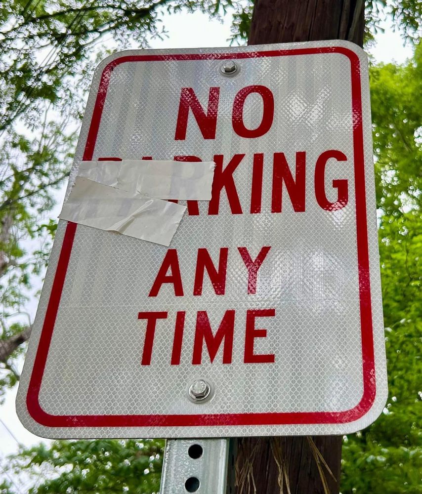 image depicts a standard “NO PARKING ANY TIME” sign that has been altered with white tape. Two pieces of tape obscure the letters “P” and “R” in the word “PARKING,” transforming it into:

”NO
—KING
ANY TIME”