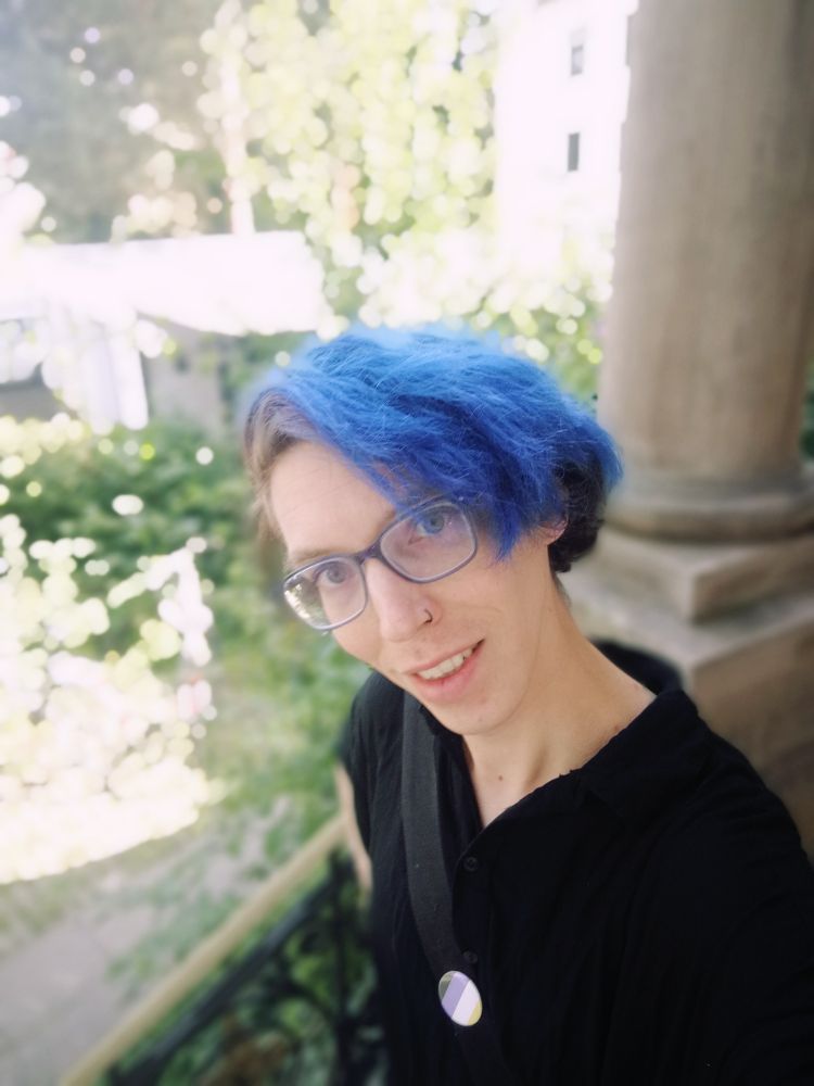 Selfie einer weißen nichtbinären Person im schwarzen Romper. Brille mit blauem Rahmen, violett gefärbte Haare, die Farbe ist zu blau verwaschen.