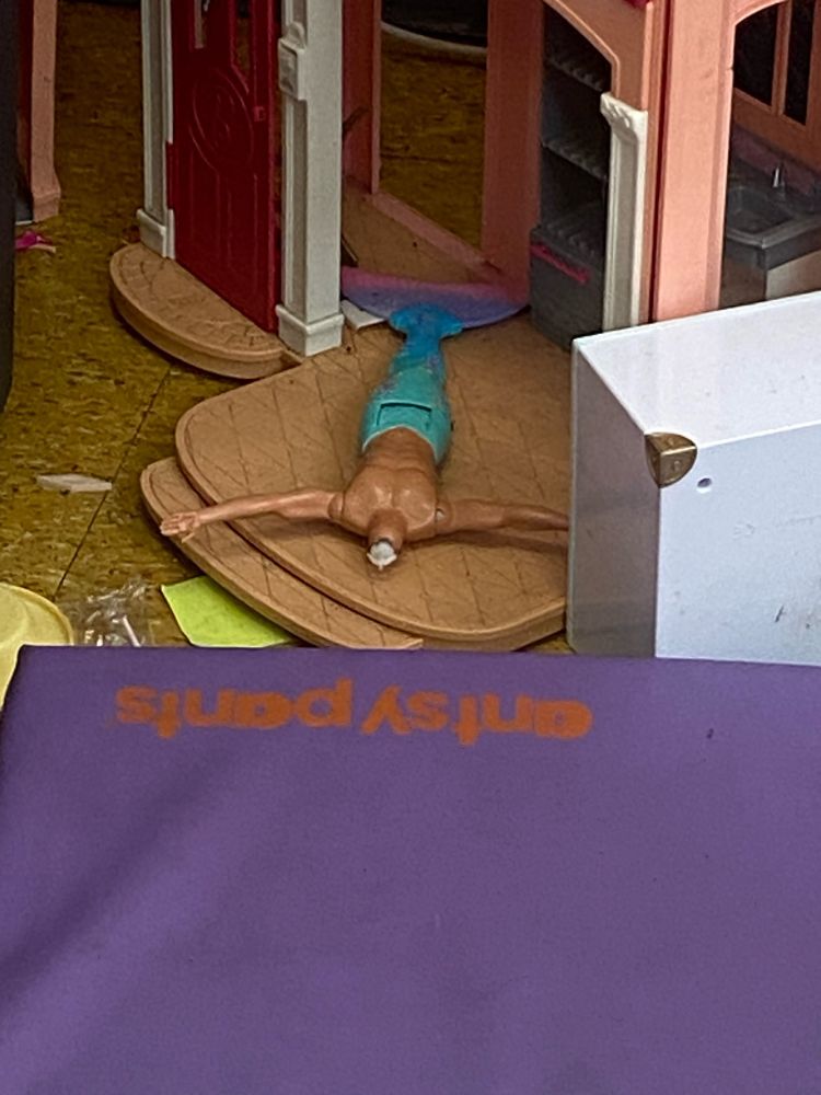 Top, a headless Barbie/Ken-proportioned merman doll lies on its back, arms outstretched, on a sort of raised landing space of a dollhouse. Along the left edge of the shot, patches of very utilitarian yellow-brown community center tile and a few scattered scraps. In the bottom foreground, a gymnastics pad prominently bisects the screen to create a field of purple, with red kiddy-font lettering near the edge (seen here upside down), reading “antsy pants.”