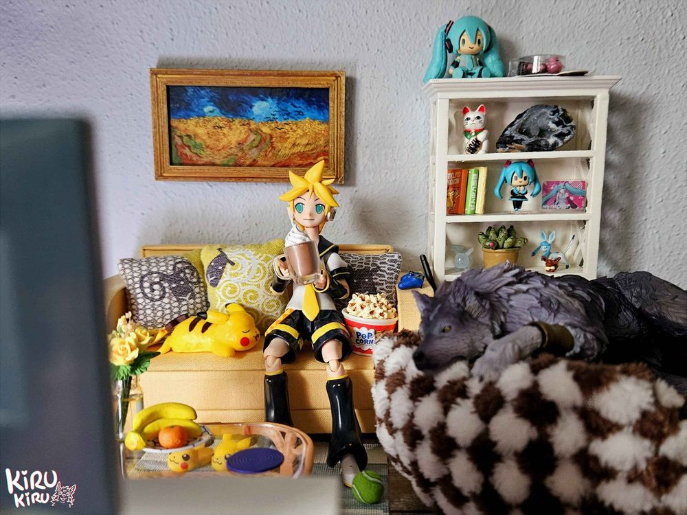 A photograph of miniatures/action figures. 
Kagamine len holding a hot chocolate while sitting on a yellow sofa with sheep pillows. On a large brown and white petbed is a wolf relaxing. They are watching tv. 
There is a shelf with small items behind them inclueding a few small mikus, a tiny eevee and glaceon, a geode, a plant and books. 
There is also a small glass coffeetable with fruit, a coaster and yellow roses in a vase. Behind it are pikachu slippers and a pikachu plush. 
On the wall is a miniature van gogh painting based on "cornfield with ravens"