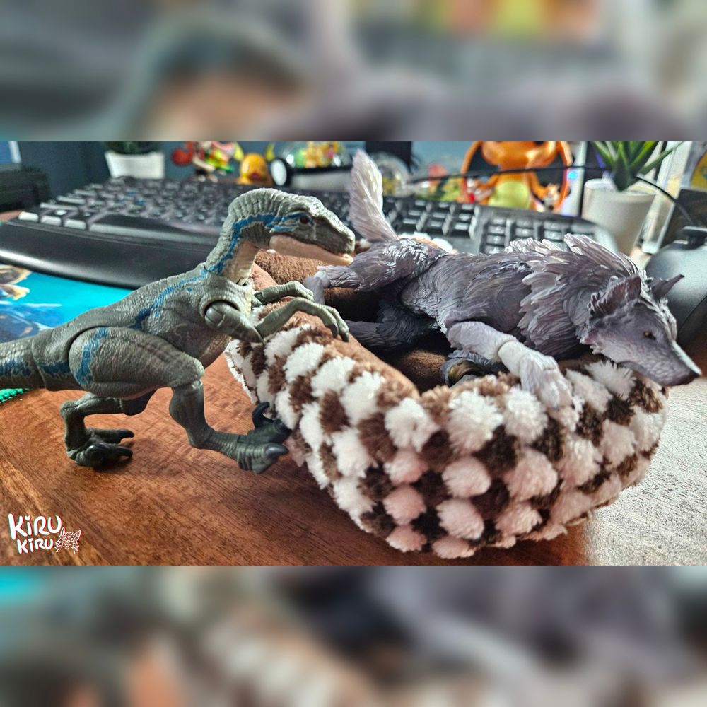 2 toy figures, a Velociraptor and a wolf.
The wolf is laying in a dogbed trying to relax while the velociraptor is trying to climb inside to join it. 
The scene is on a desk, one  an see toys, a keyboard and a controller among other things in the background