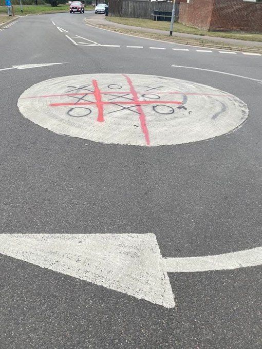 Photo of a small white circle denoting a roundabout with a red cross on it where someone has drawn a second red cross and then filled in the squares with noughts and crosses. UK