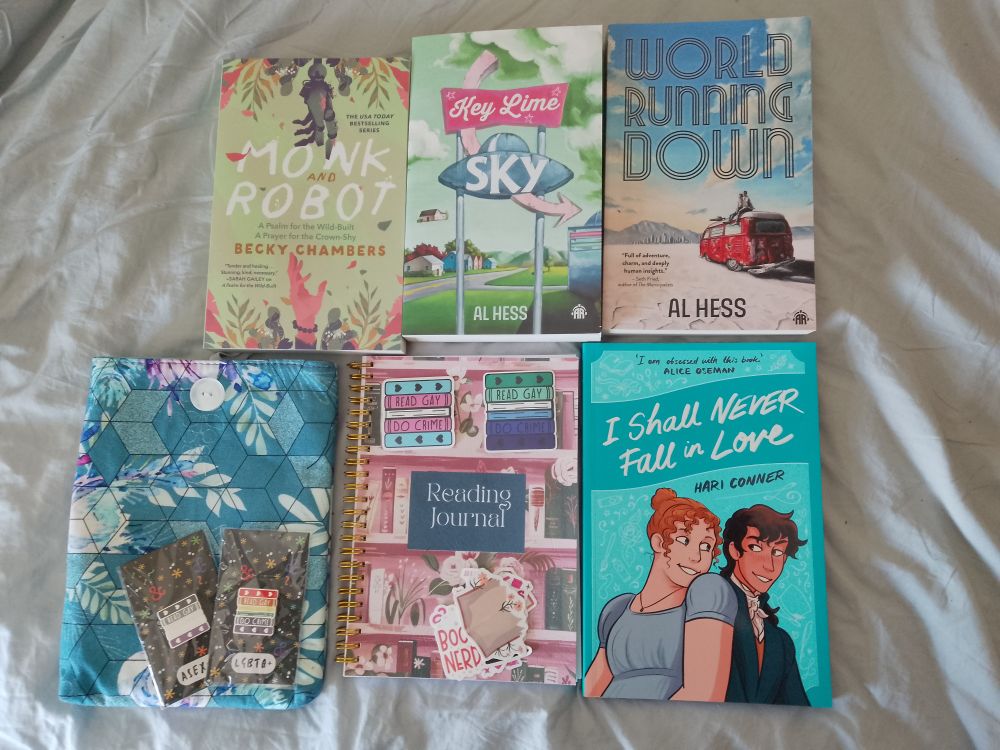A photo of four books and other book-related goodies, including stickers and pins that read 'read gay do crime', a reading journal and a blue book sleeve. The books are I Shall Never Fall in Love by Hari Conner, Monk and Robot by Becky Chambers, and Key Lime Sky and World Running Down by Al Hess