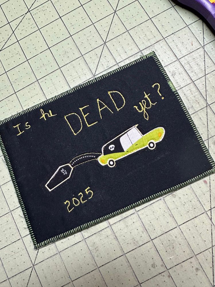 Black fabric postcard with a hearse with a coffin falling out of the back and the words “is he dead yet?” Stitched onto it. 
