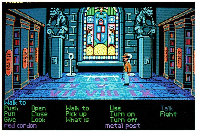 An early screenshot of Indiana Jones and the Last Crusade adventure game, from Game Player's PC Strategy Guide vol. 2 #4, September/October 1989. Indy is standing in the Venice library near a stained glass window, facing left. His whip is visible on his sprite, being held in his right hand. Instead of the "Travel" verb, a "Fight" verb, which is green as if it might be used, appears in the GUI instead.