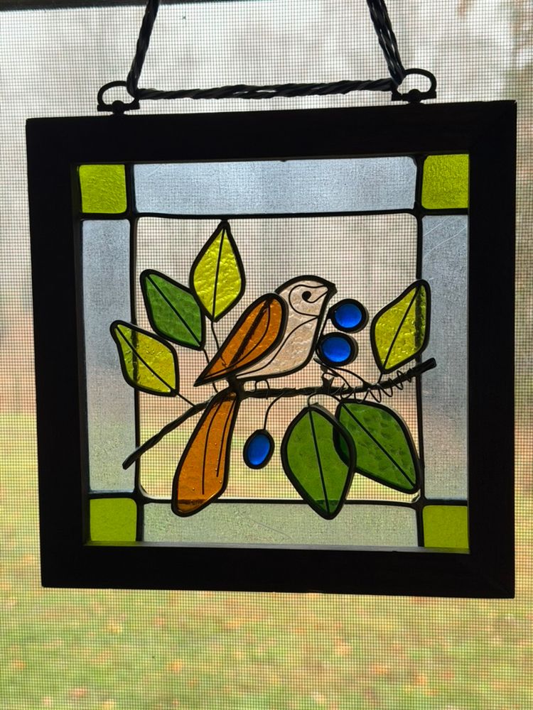 Stained glass of a small bird with brown wing and tail sitting on a branch with two shades of green leaves and blue berries. It hangs in my "bluebird" room named after the shade of paint on the walls. Old. A gift to my grandmother in the 1970's and returned to me after her passing many years ago.