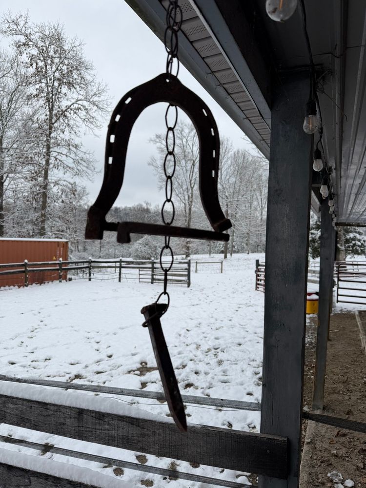 Country dinner bell made from two extra large horse shoes welded together with a railroad spike hanging from a chain to use as a striker. It hangs on the barn porch with a snowy December 1, 2025 paddock and pasture behind.