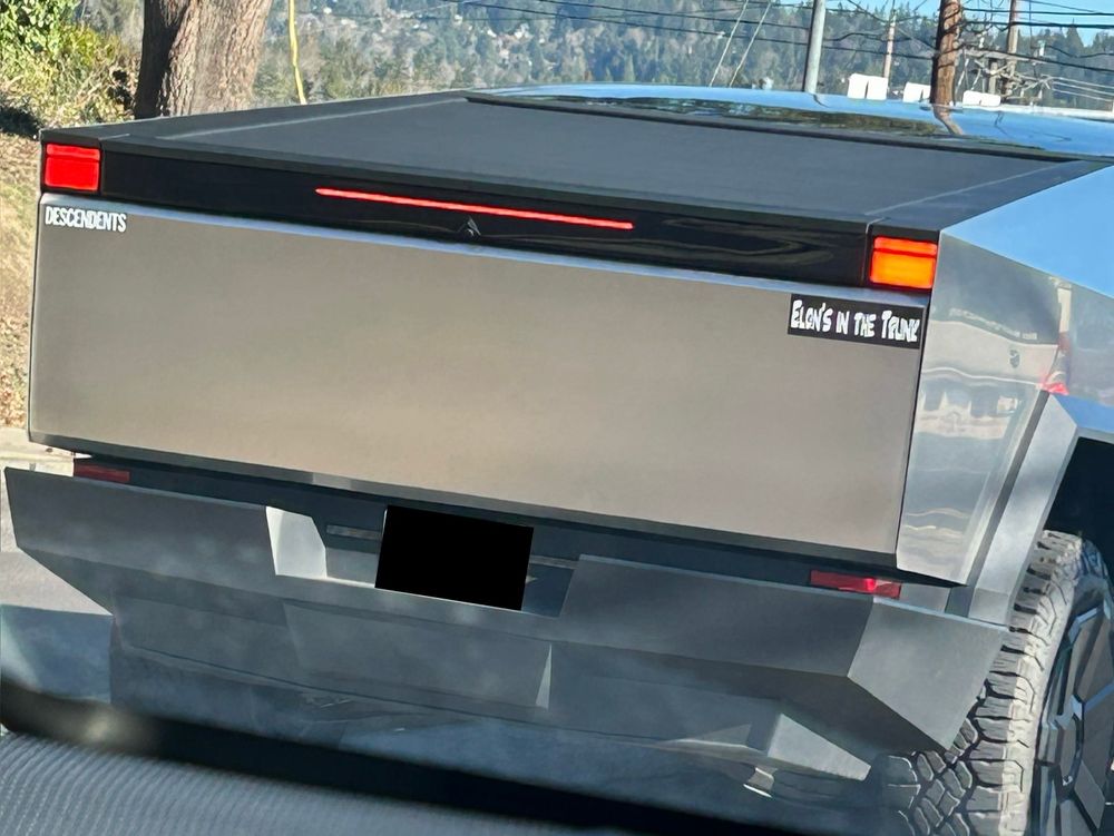Rear of a cybertruck with a Descendants (late 70s punk band from SoCal) bumper sticker on one side and an “Elon’s in the trunk” sticker on the other. 
