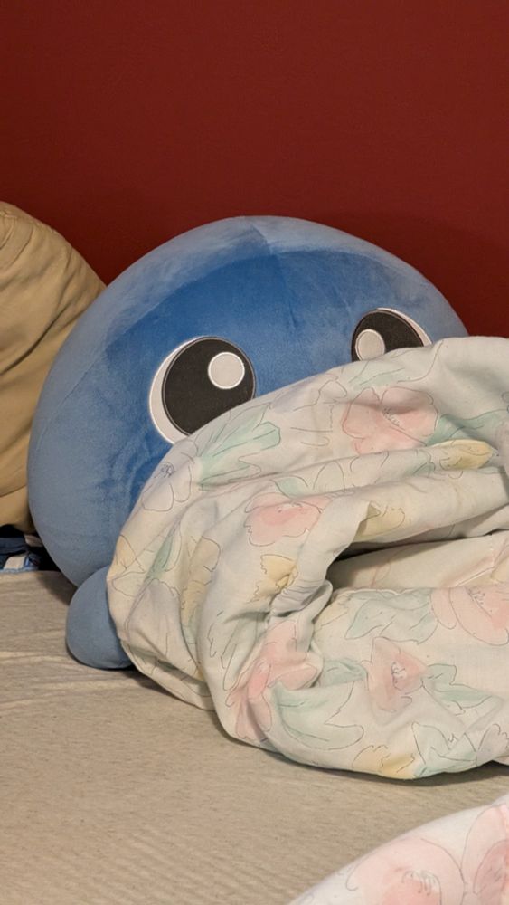 Giant Poliwag plush on a bed. It's half covered by a blanket so it looks like it's peeking out.