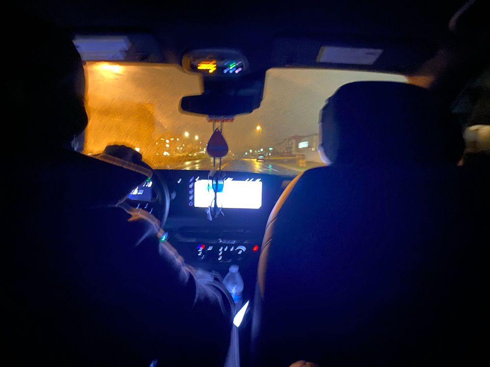 Picture of a man driving at night taken from the back seat. The street is black with water and the light outside is very pronounced