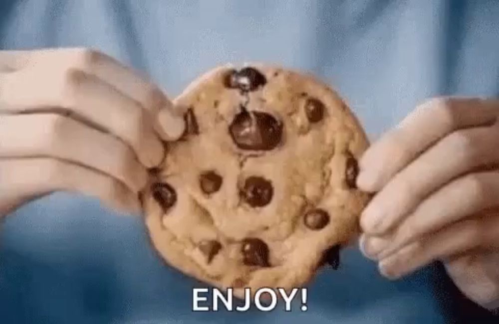 a person is holding a chocolate chip cookie in their hands and says `` enjoy '' .