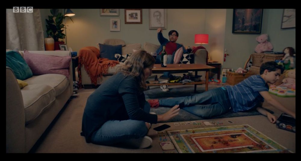 A screencap from the TV show There She Goes, series 1 episode 1. Simon Yates is sat on the sofa, whilst his wife Emily and son Ben play Talisman on the living room floor. 