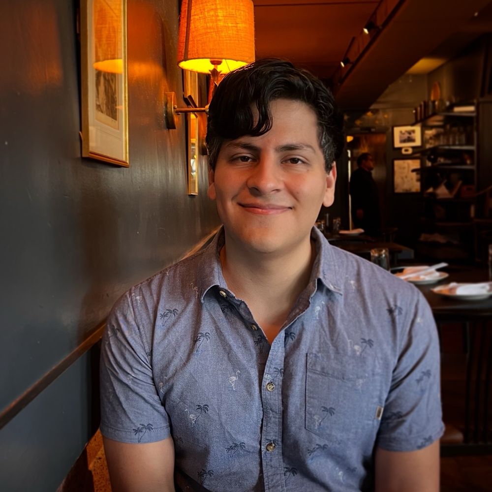 Smiling young man in short sleeved button up facing the camera in a warmly lit restaurant 