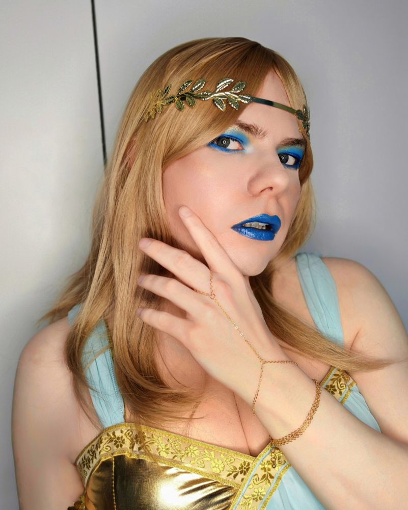 Aly with blonde hair and blue makeup in a white, gold and light blue ancient greek style dress with various gold accessories. This shot in particular shows off a gold bracelet with a chain up to my middle finger.