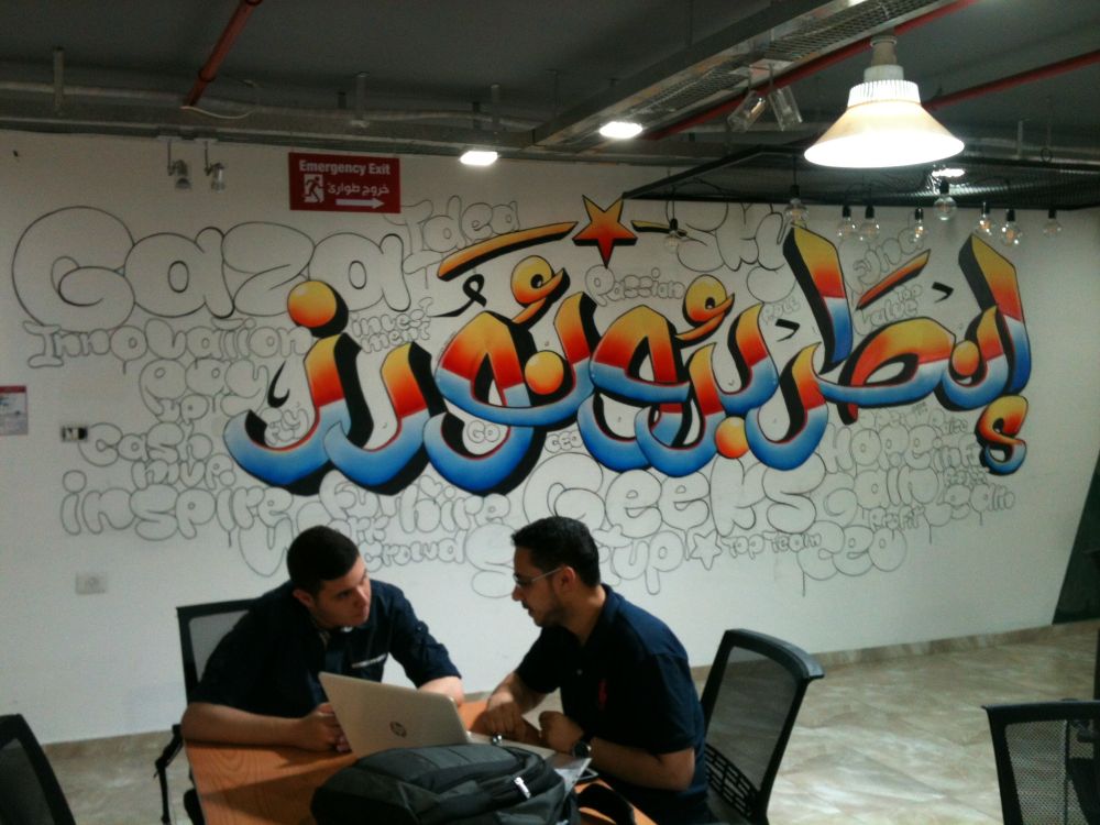 Blue and orange Arabic script graffiti on a white wall covered in English cartoon outlined English words like "Gaza", "Innovation", "Inspire", "Geeks", "Hope," "Idea", "Pay up", "Fly", "MVP", etc. In the foreground, two 20-something men sit working together on a laptop in front of them on a table. 