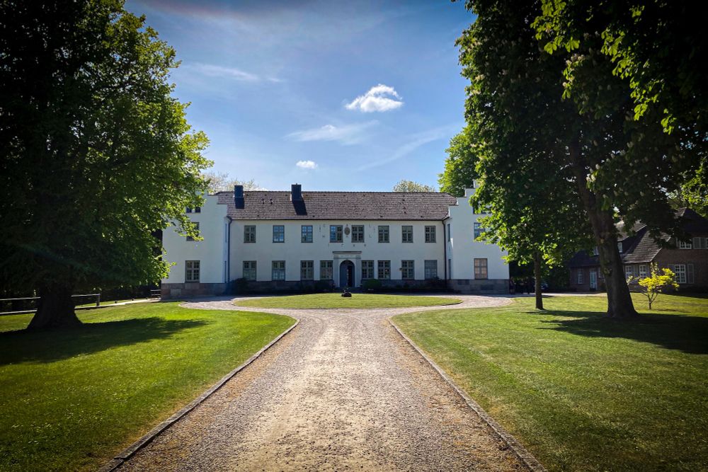 Ein zweistöckiges weißes Landhaus mittig im Bild, eingerahmt von Laubbäumen