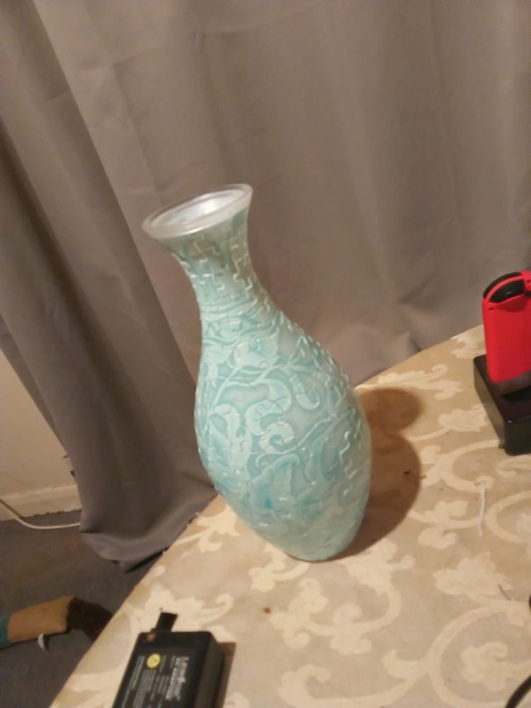 A completed 3d puzzle vase with a pale blue floral design at a jaunty angle on a table whose cloth has a yellow floral design. Behind as a backdrop are grey curtains
