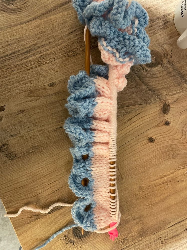 Knitting on a knitting needle. The edge is pale blue scallops and there is one pale pink stripe in stocking stitch 