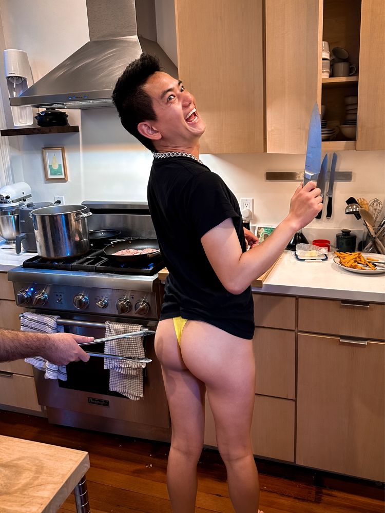 Me making an exaggerated chaotic face. I’m holding a knife and in a yellow thong in the kitchen. Butt cheeks exposed. An arm is coming from left side of frame holding a clamper thing (tongs?) towards my butt