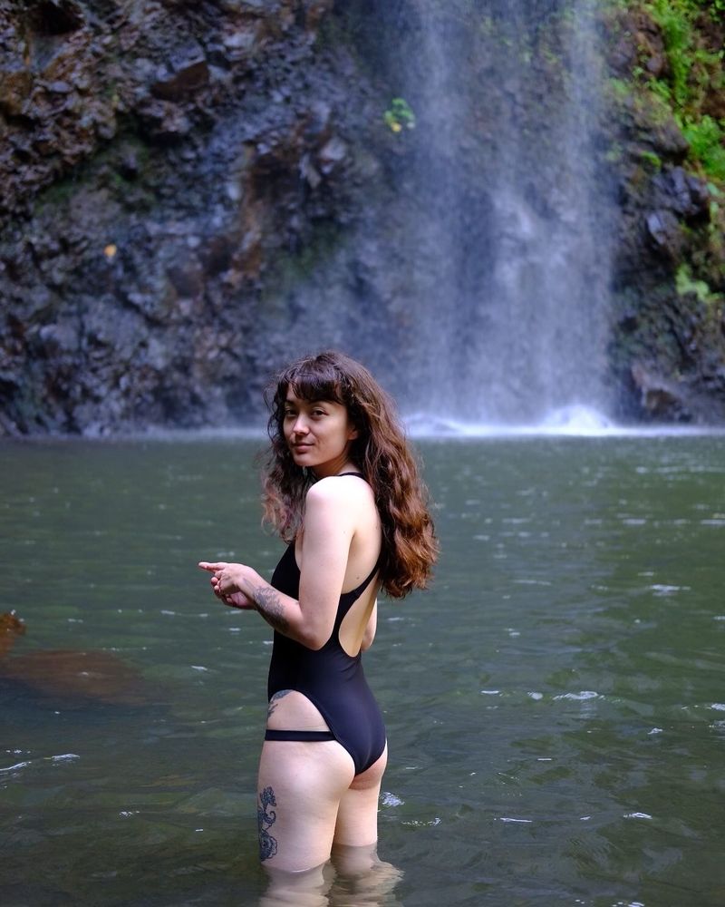 Juno, a half-Japanese woman with long wavy brown hair, wearing a one piece black bathing suit stands in a pool of water beneath a beautiful waterfall.