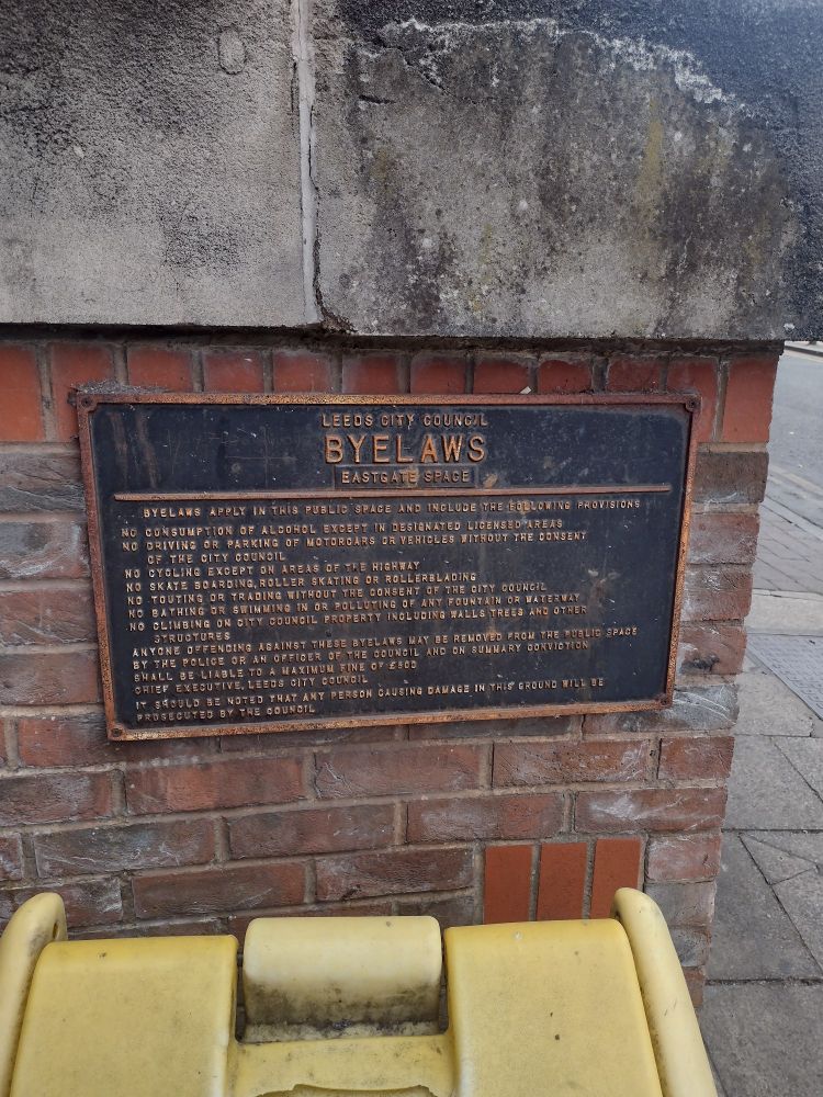 A public notice describing in detail byelaws of the Leeds Eastgate Space. The sign looks to be quite old given the raised lettering on the sign has succumbed to rusting and now has a dirty orange look to it. 