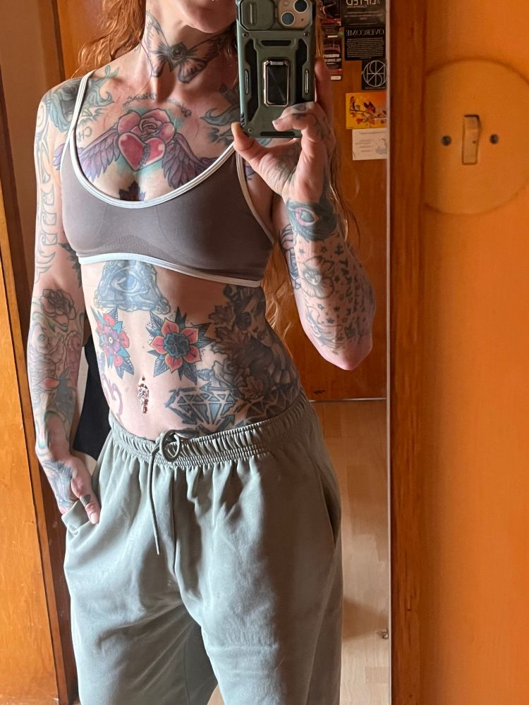 Selfie einer bunt tätowierten Frau mit khakifarbener Jogginghose und Mokkafarbenem BH mit weißem Rand 