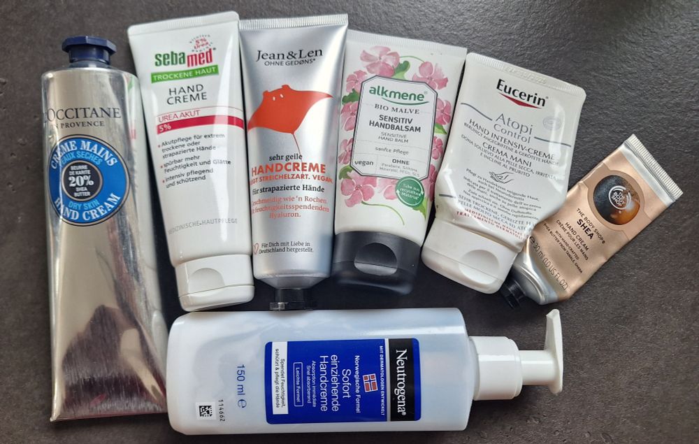 7 Handcremes liegen auf einer Küchenabeitsplatte. Es handelt sich um L'Occitane Karité, Sebastian Med Urea Akut, Jean&Len Sehr geile Handcreme (die mit dem Rochen drauf), Alkmene Bio Malve Sensitiv Handbalsam, Eucerin Atopi Control Hand Intensiv-Creme, The Body Shop Shea Hand Cream und Neutrogena Sofort einziehende Handcreme. Alles in Tuben, außer letztere in einem Pumpspender.