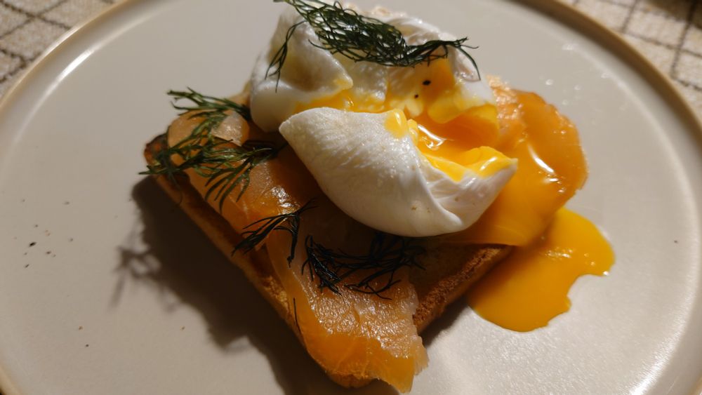 Ein pochiertes Ei, bei dem der noch flüssige Dotter ausläuft, auf einem Toast mit Dillsenf, Räucherlachs und Dill.