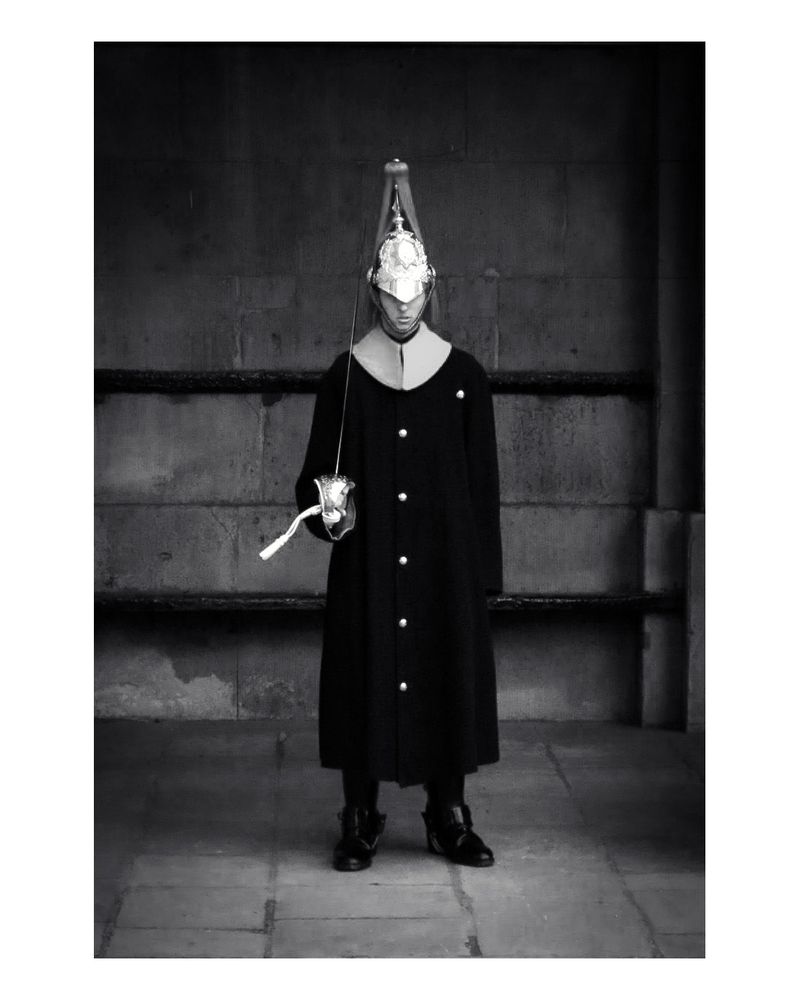 A black and white photo of a member of the King's Troop Royal Horse Artillery in full dress uniform. They are wearing a helmet with a plume, a long coat with white buttons and collar, and holding a sword in their right hand. The background is a dark stone wall.