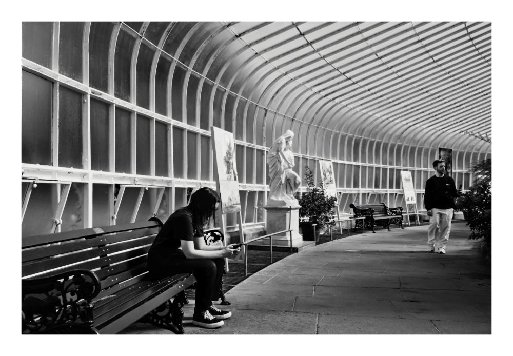 Black and white photograph inside a Victorian-style glass and iron conservatory. A person with long, dark hair wearing dark clothes and high-top sneakers sits on a wooden bench in the left foreground, looking down at a mobile phone. In the center, further back, stands a classical white marble statue of a draped female figure on a pedestal. To the right, a man in a dark-colored top and light trousers walks towards the viewer along the curved stone floor. The roof is a dramatic curved, segmented structure of glass and white metal ribs, creating a strong linear perspective and high-contrast light and shadow.