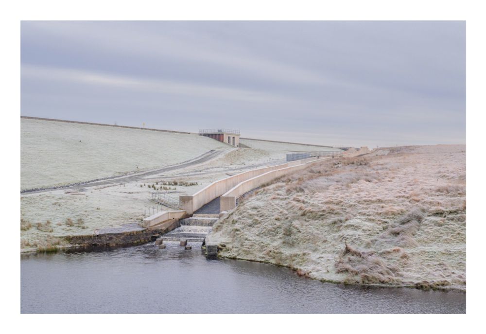A concrete dam and spillway stretch across a frozen lake. The lake is covered in a thin layer of ice and snow, and the surrounding landscape is also covered in frost. The sky is overcast and grey.