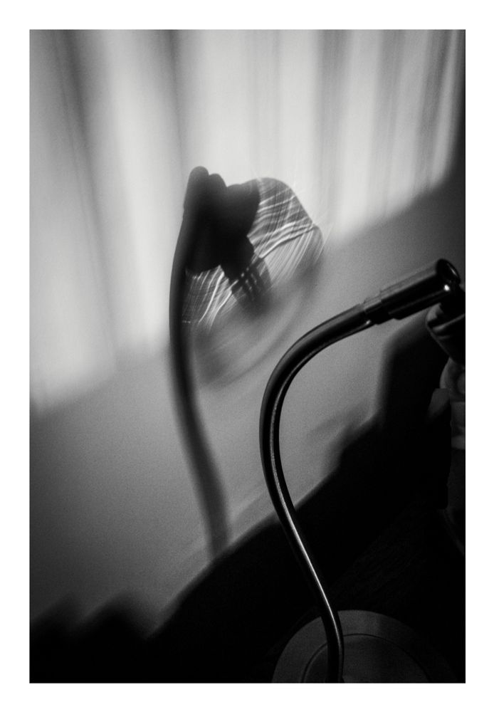 A high-contrast, black and white photograph of a flexible-necked desk lamp. The lamp is partially visible in the bottom right corner, with its curved metallic neck leading up. The light source is out of frame but casts a strong, overexposed, and blurred shadow onto the textured wall behind it. The shadow is elongated and features distinct, sharp streaks of light emanating from the lamp's head. The overall mood is moody and abstract, emphasizing light, shadow, and motion.