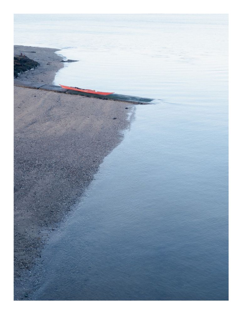 A red kayak rests on a sandy beach, angled towards the water's edge. The water is calm and clear, with a gentle gradient from light blue to dark blue. The beach is pebbled, and a small concrete structure extends into the water. The sky is overcast and pale.