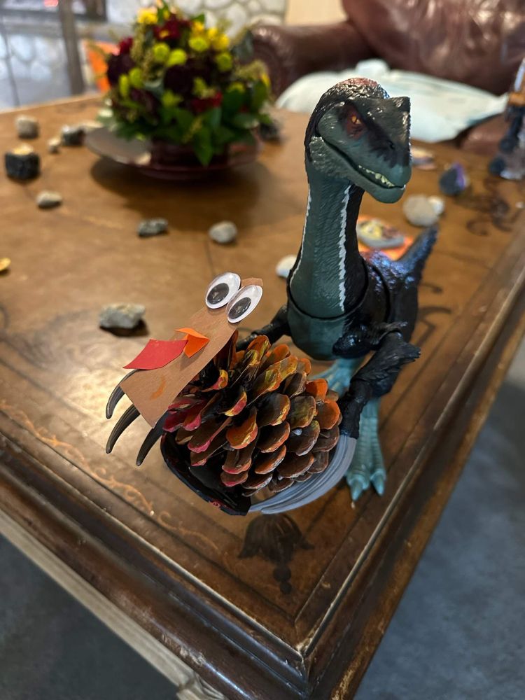 A plastic dinosaur holding a pinecone made to look like a turkey with colored paper and googly eyes.